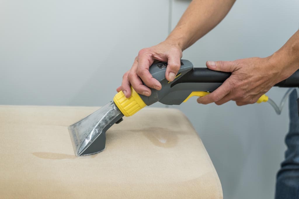 Eine Person verwendet eine handgeführte Polsterreinigungsmaschine mit einem gelben und grauen Griff, um eine hellbeige Stoffoberfläche zu reinigen.