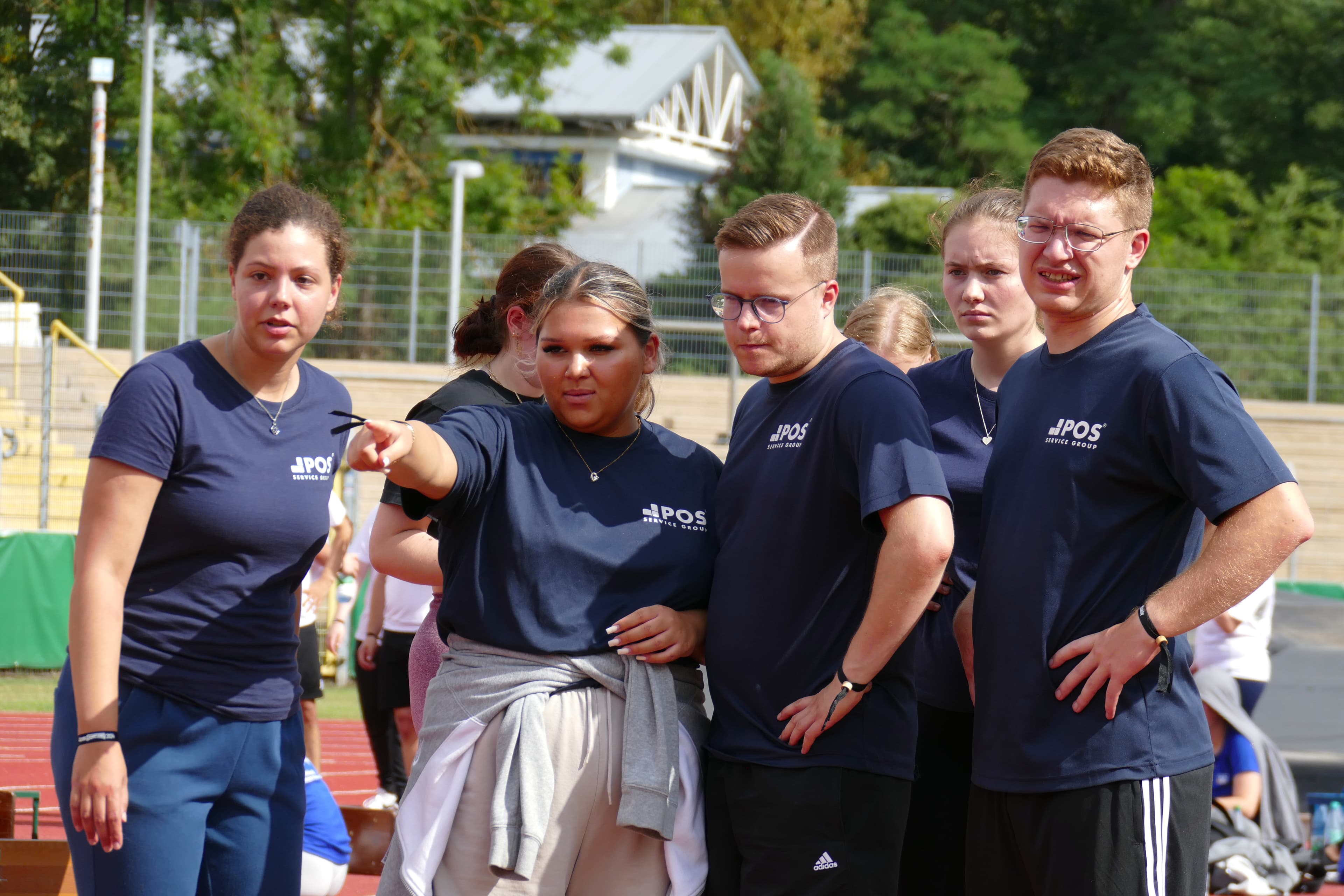 Vijf jongvolwassenen in bijpassende marinehemden staan samen buiten op een sportbaan. Eén vrouw wijst naar iets buiten het kader terwijl de anderen in dezelfde richting kijken met gefocuste of bezorgde uitdrukkingen.