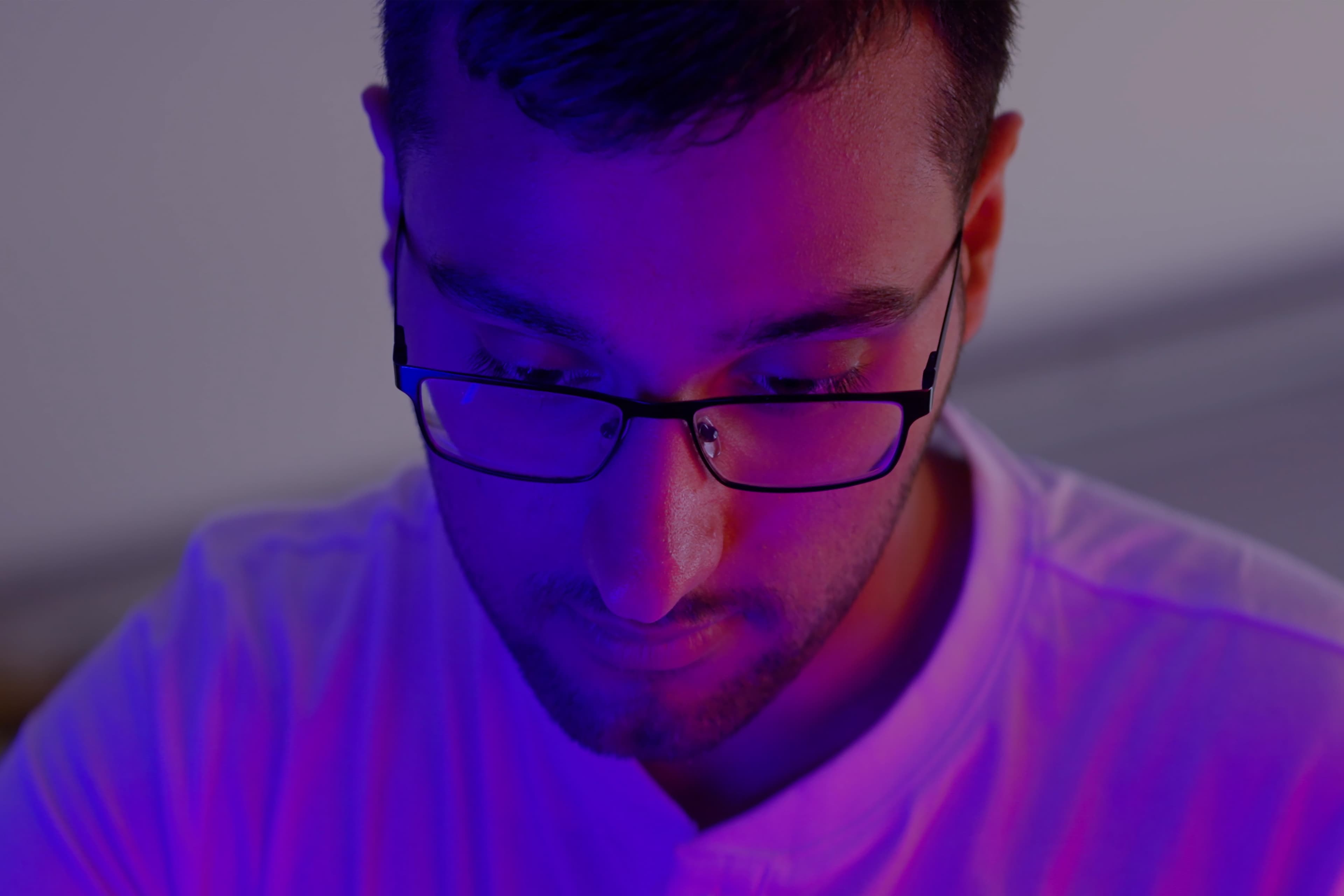 Een persoon met een bril en een wit overhemd kijkt naar beneden, verlicht door paarse en blauwe verlichting in een schemerige kamer.