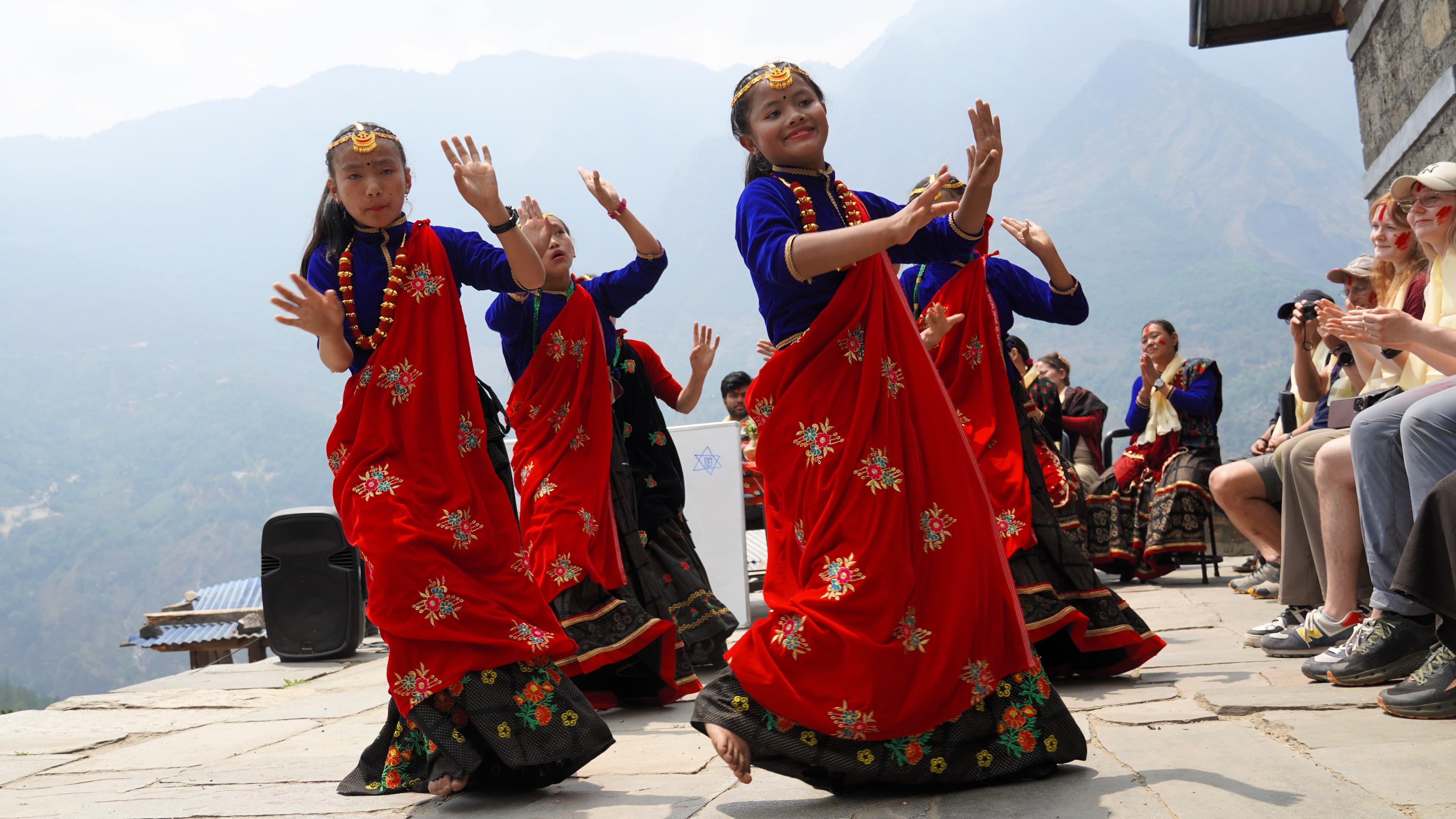 Eine Gruppe von Frauen in leuchtend roten und blauen traditionellen Kleidern führt im Freien einen kulturellen Tanz auf, mit Bergen im Hintergrund und Menschen, die von der Seite zusehen.