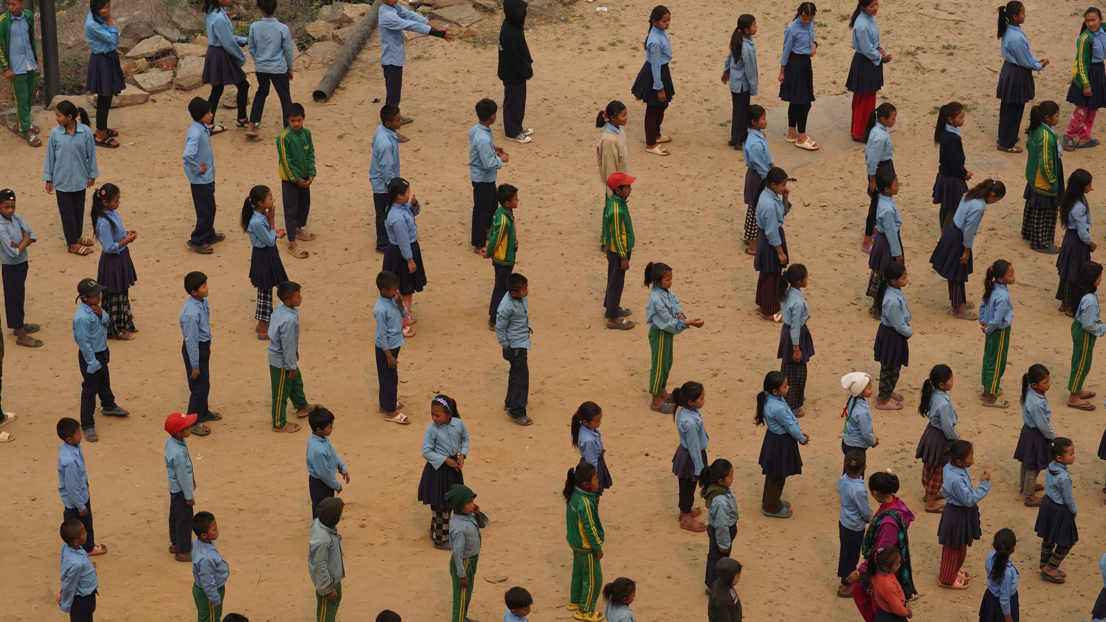 Eine große Gruppe von Schulkindern, die blaue Hemden und grüne Trainingsanzüge tragen, stehen in ordentlichen Reihen auf einem sandigen Boden und nehmen wahrscheinlich an einer Schulversammlung oder einer Aktivität im Freien teil.