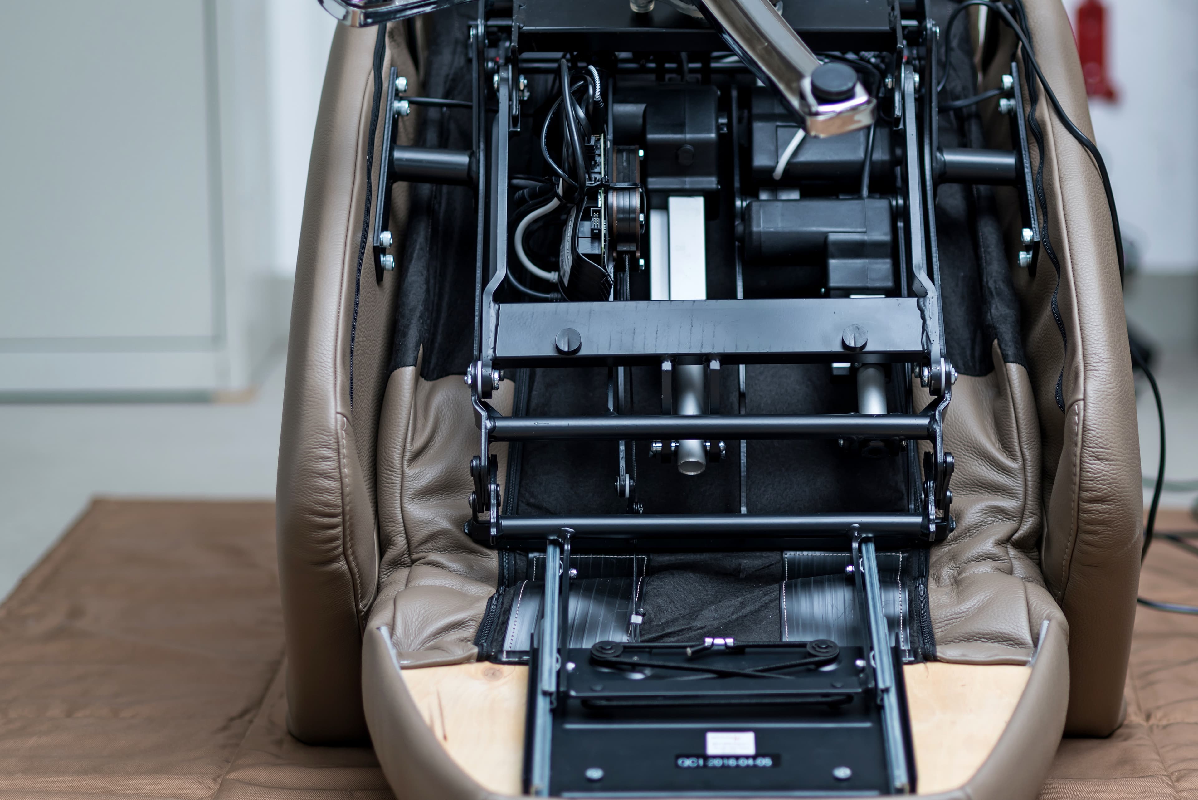 Das Bild zeigt die Unterseite eines braunen Lederlehnstuhls, wobei die mechanischen und elektrischen Komponenten, einschließlich Metallrahmen, Drähte und bewegliche Teile, sichtbar werden. Die Polsterung ist teilweise geöffnet und gibt den Blick auf das Innere frei.