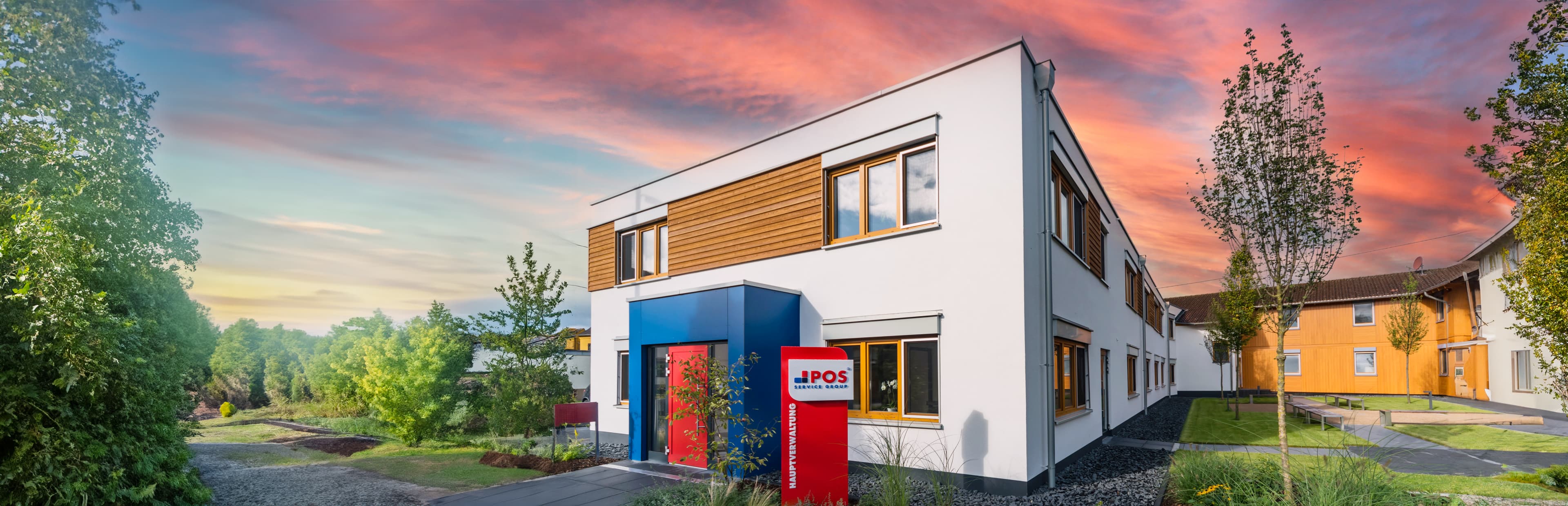 A modern two-story building with white and wood paneling sits amid greenery at sunset, with a red sign labeled IPOS near the entrance and a colorful sky in the background.