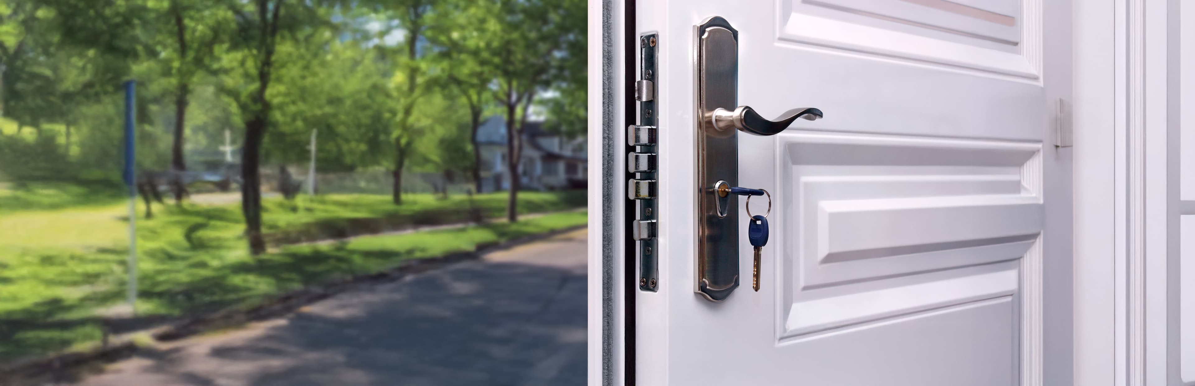 A white door with a modern handle is slightly open, revealing a key inserted in the lock. Outside, there is a sunny neighborhood street lined with trees and green grass.