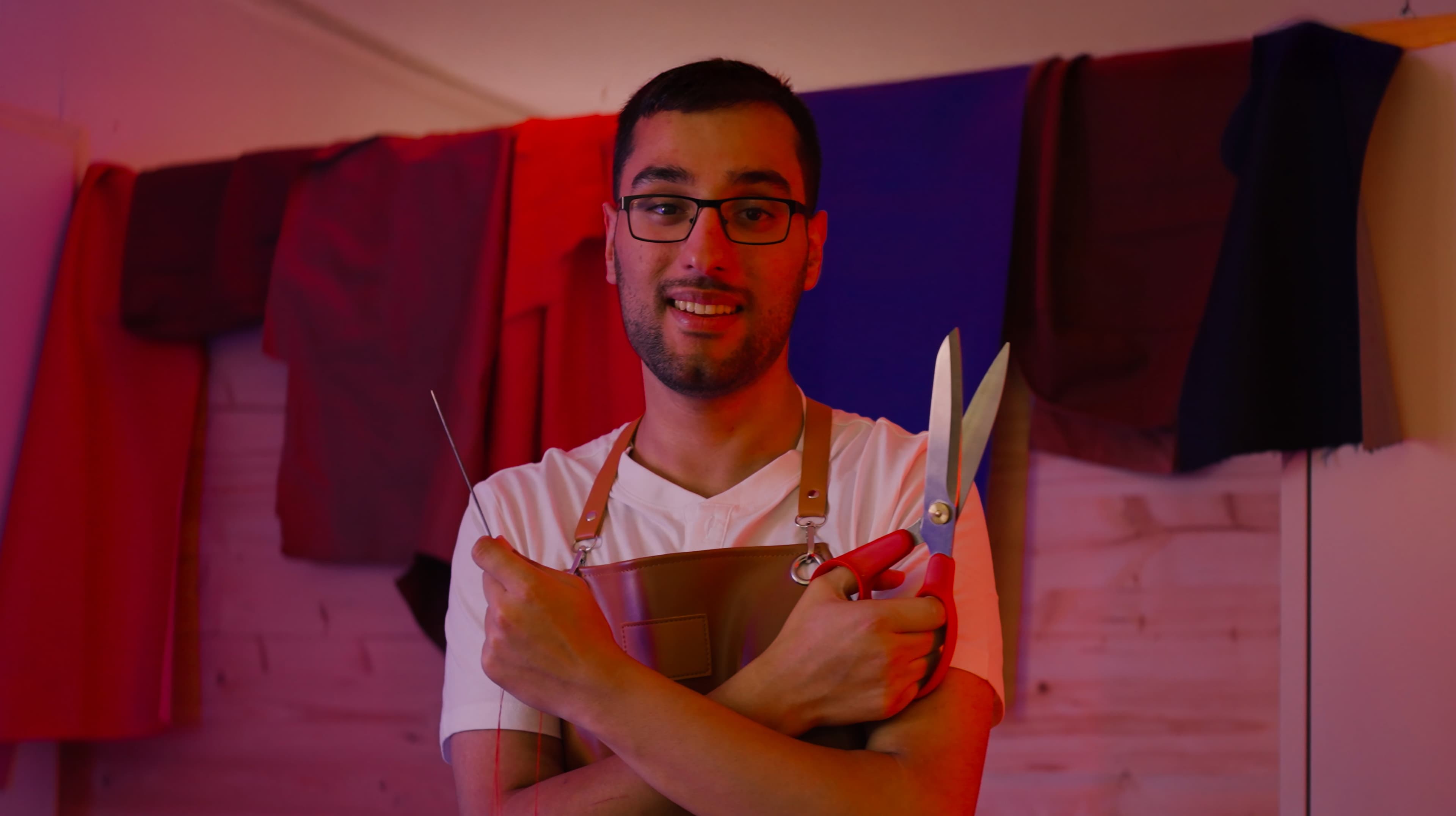 A man wearing glasses and an apron stands in front of colorful draped fabrics, smiling and holding large scissors and a tool in his crossed arms.