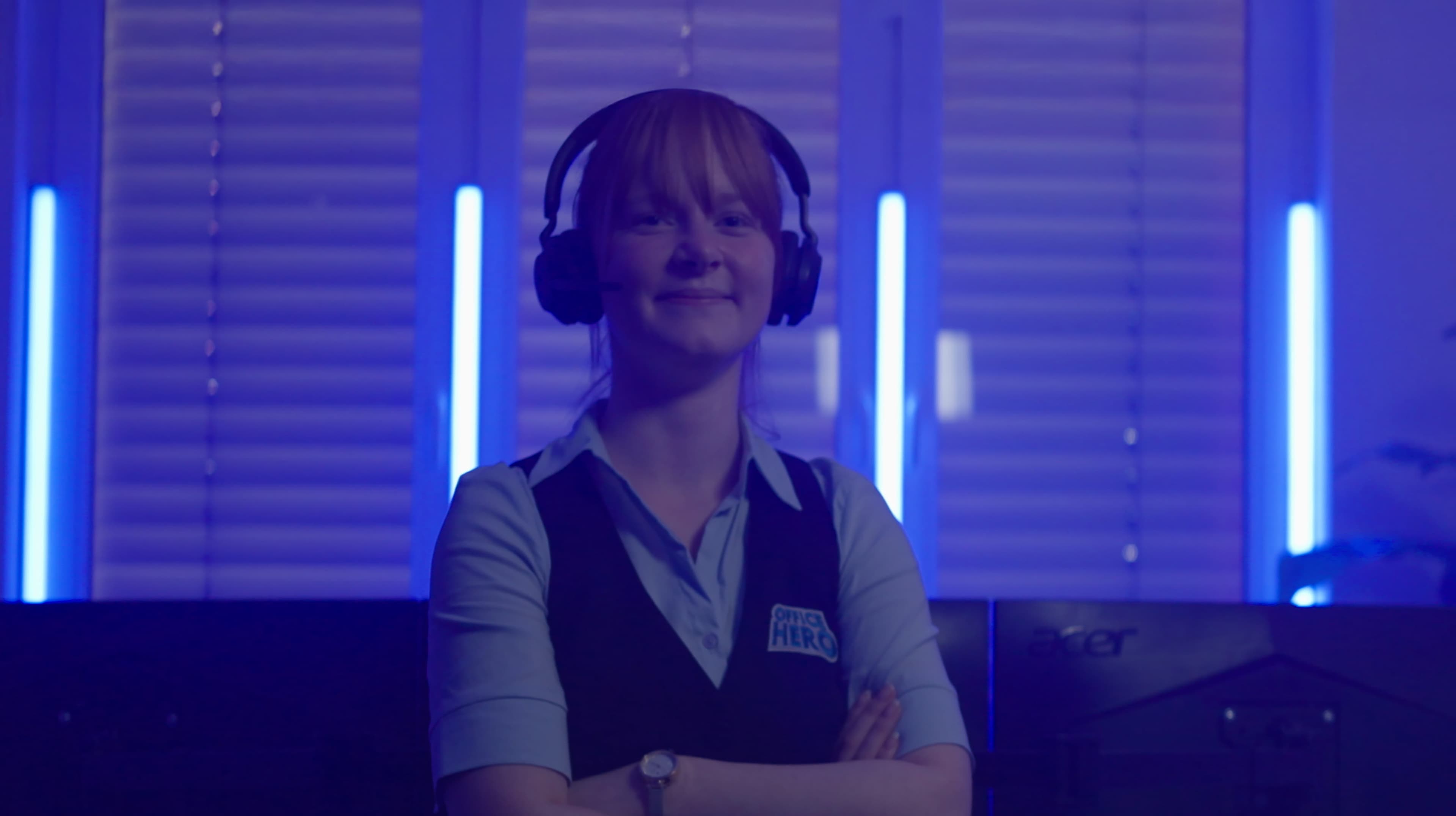 A smiling woman wearing a headset and a blue vest with a HIRE HERO badge stands with arms crossed in a dimly lit room with blue lights and computer monitors.