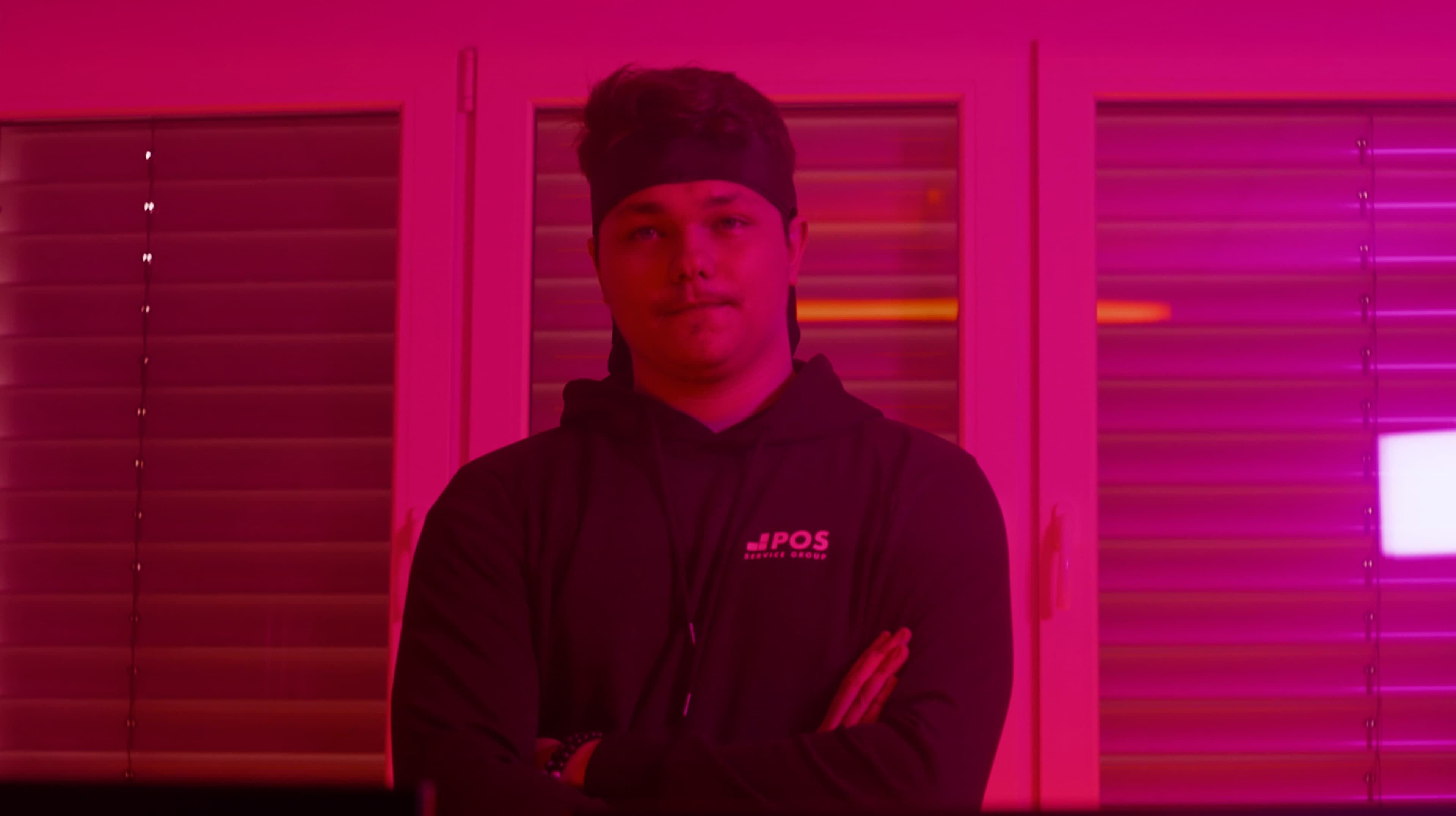 A person wearing a headband and a dark hoodie stands with arms crossed in front of closed blinds, illuminated by pink-purple lighting.