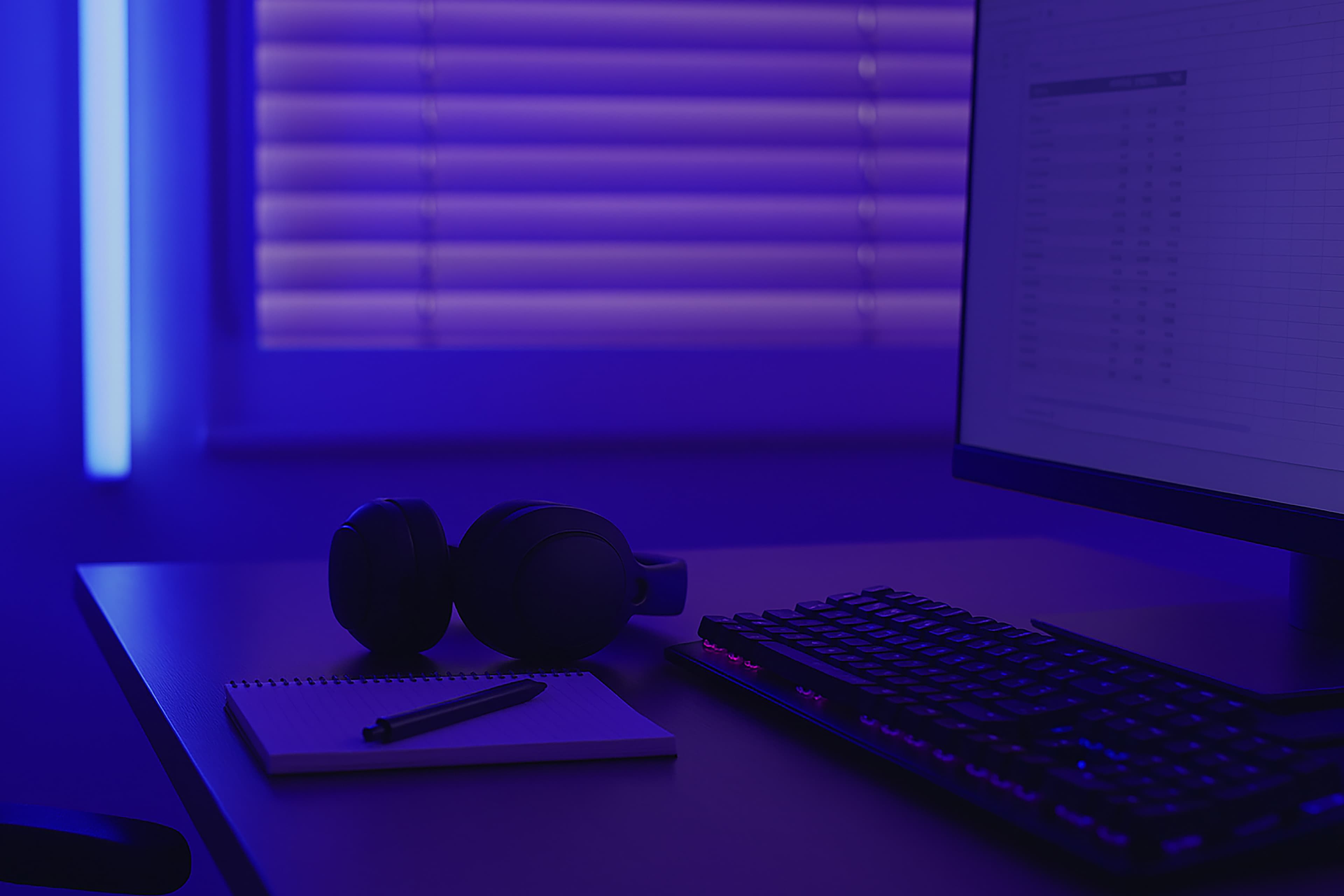 Een bureau met een toetsenbord, computermonitor, notitieblok, pen en koptelefoon, verlicht door paarsblauwe omgevingsverlichting van een raam met gesloten jaloezieën op de achtergrond.