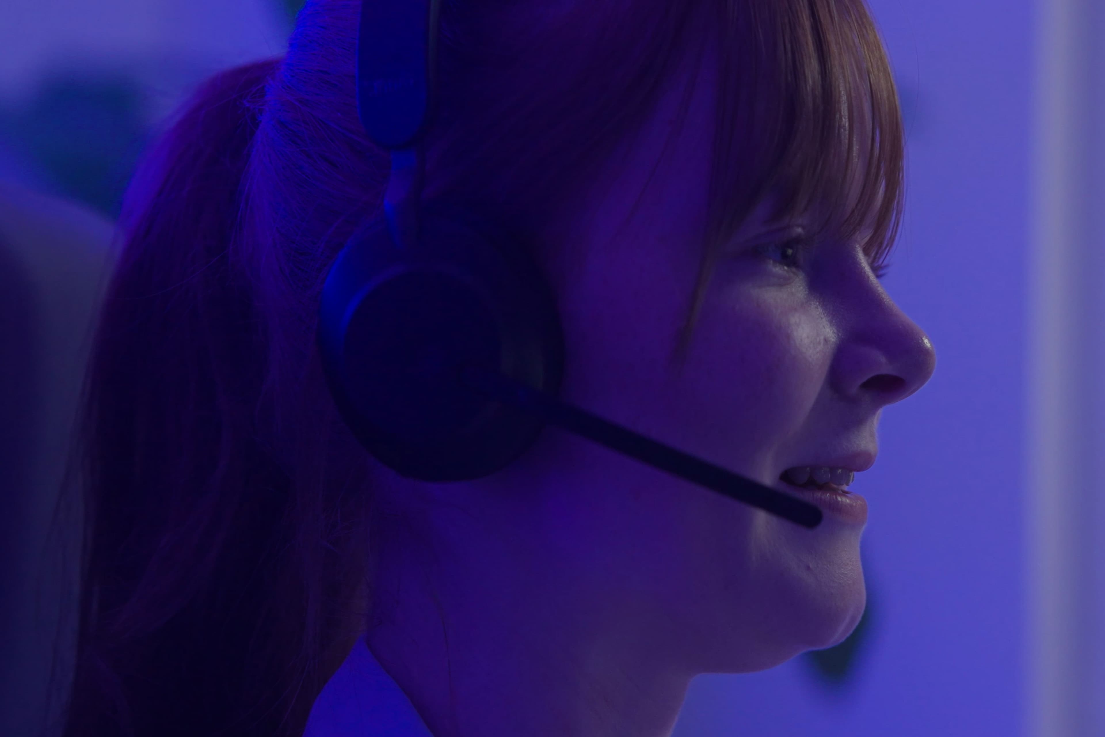Een close-up van een lachende persoon met rood haar die een headset-microfoon draagt, badend in blauw licht, wat suggereert dat ze bezig zijn met een gesprek of aan het gamen zijn.
