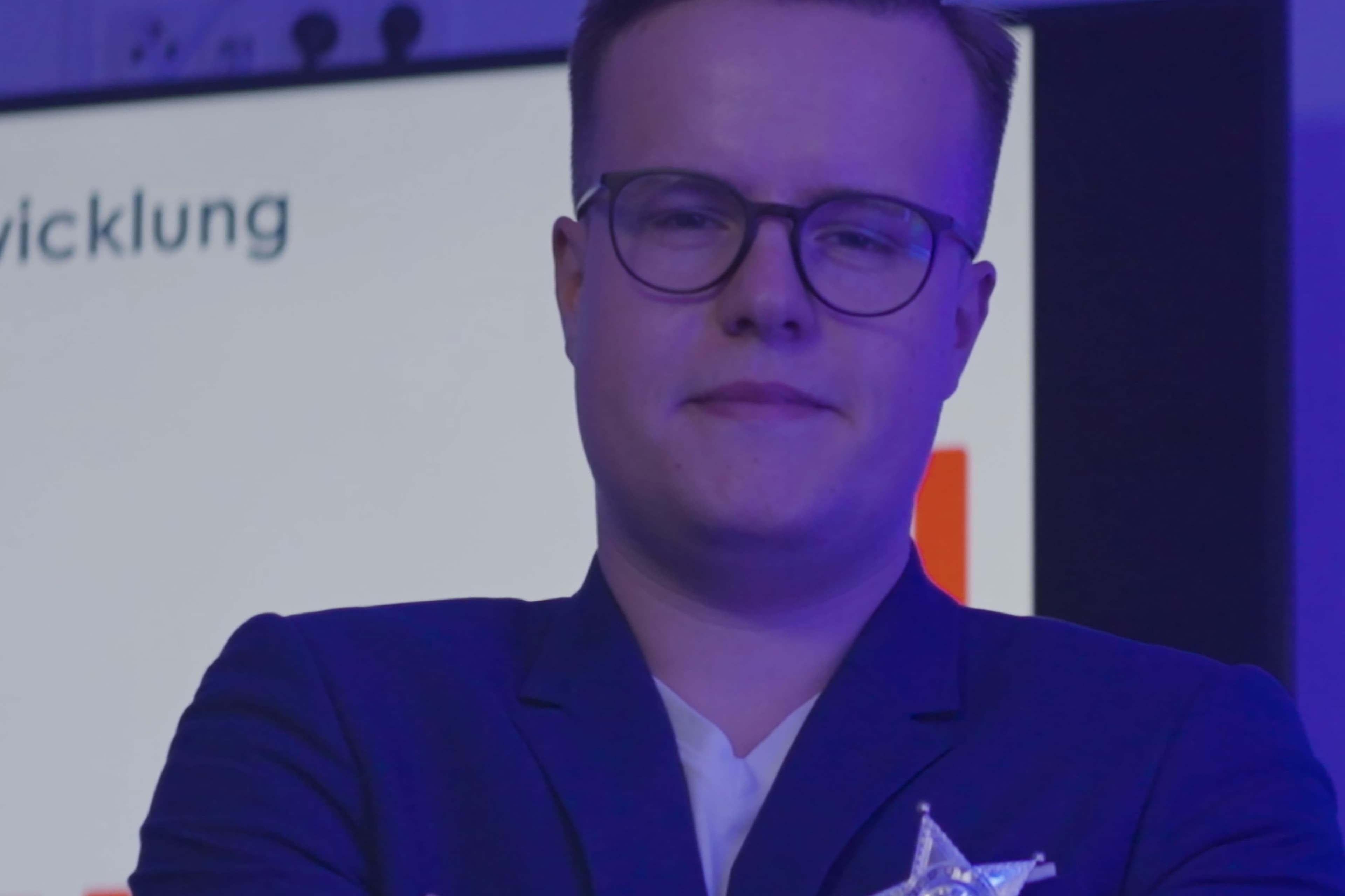 A person with short hair and glasses stands in front of a blurred presentation screen, wearing a dark blazer and white shirt, with purple-blue lighting.