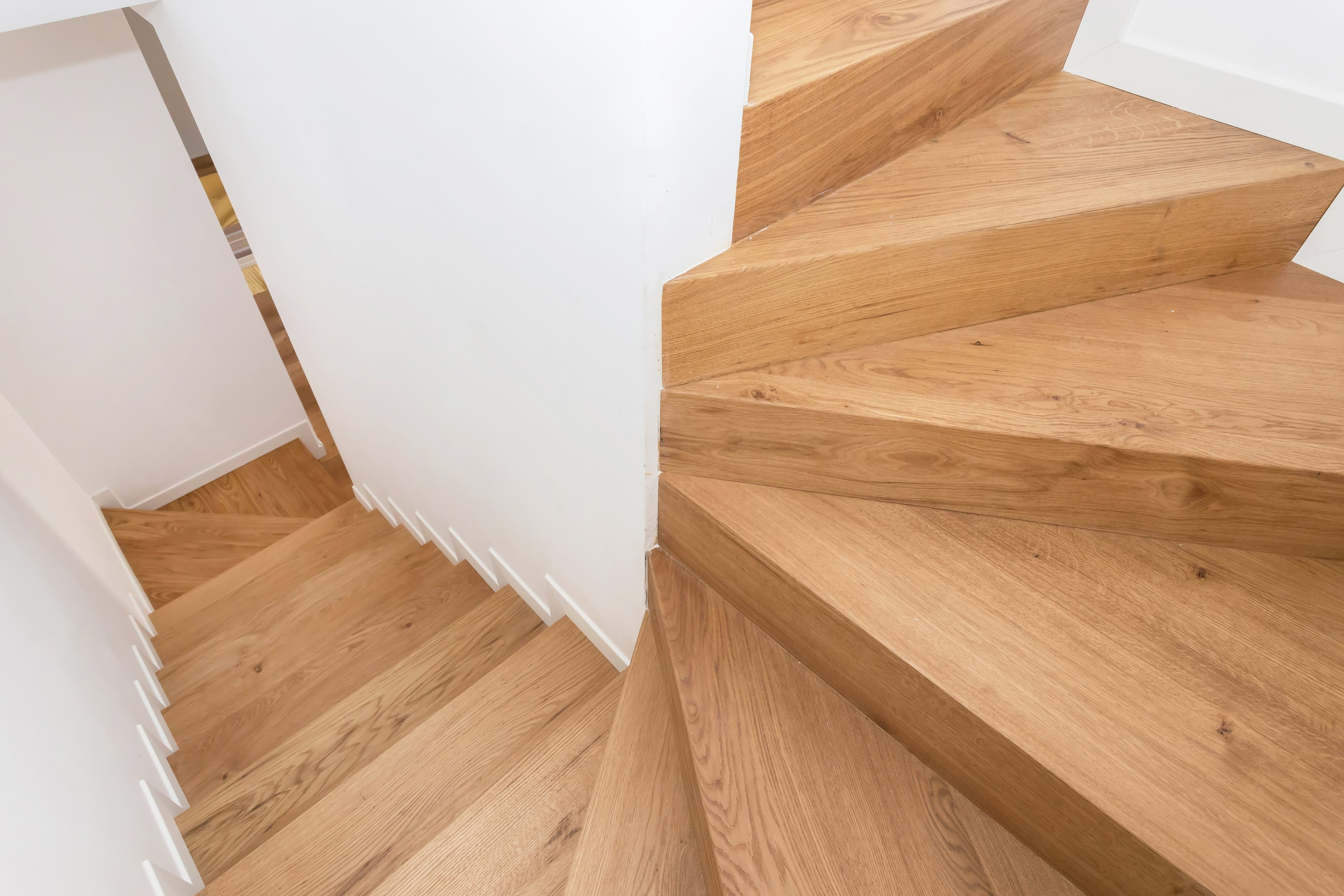 Vue rapprochée d'un escalier moderne en bois avec des marches marron clair et des murs blancs, caractérisé par un virage serré et des angles géométriques. L'escalier est d'un design épuré et minimaliste.