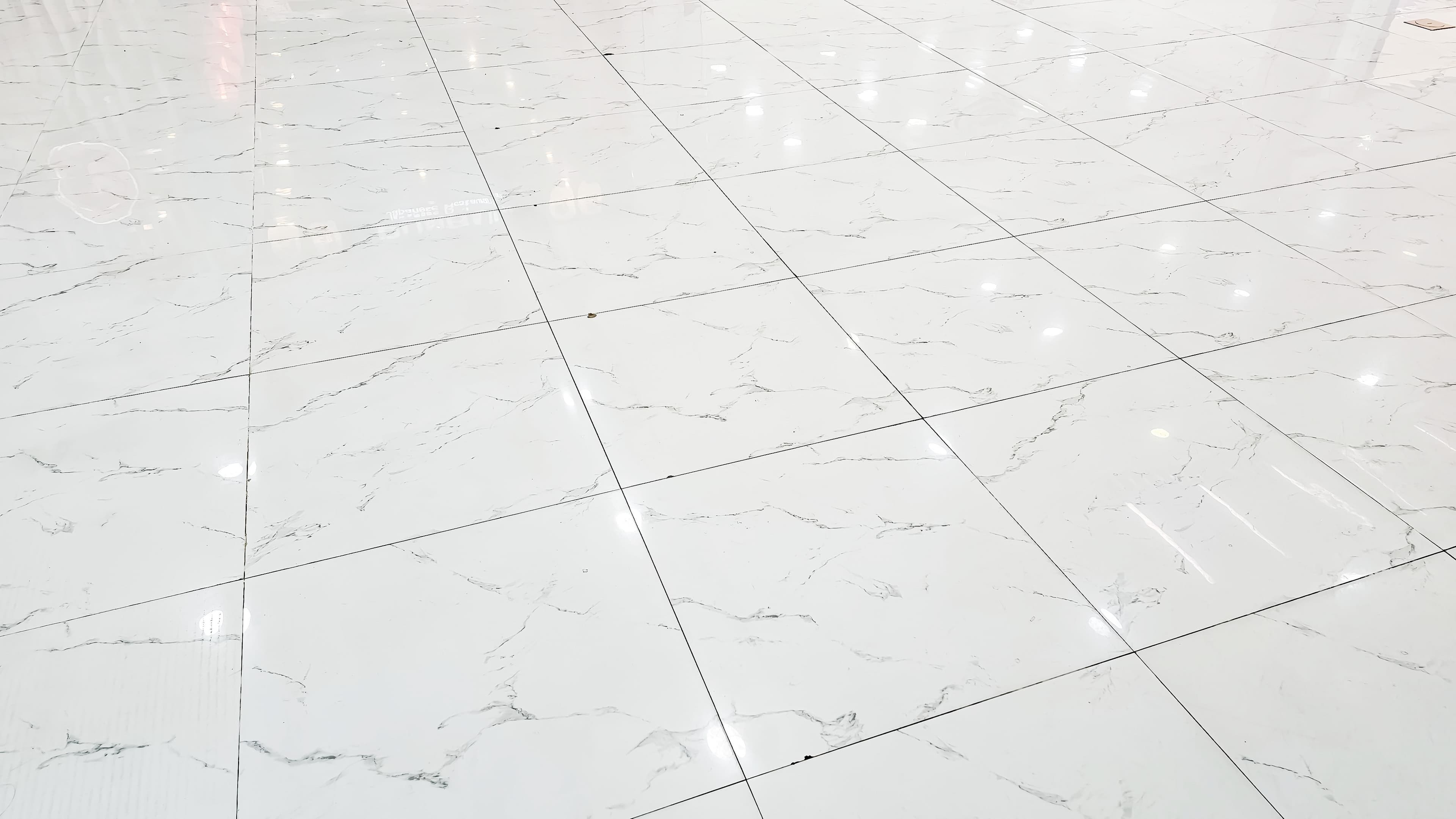 Un sol en marbre blanc brillant avec de légères veines grises et des lignes de coulis noires en forme de grille, reflétant les lumières vives du plafond.