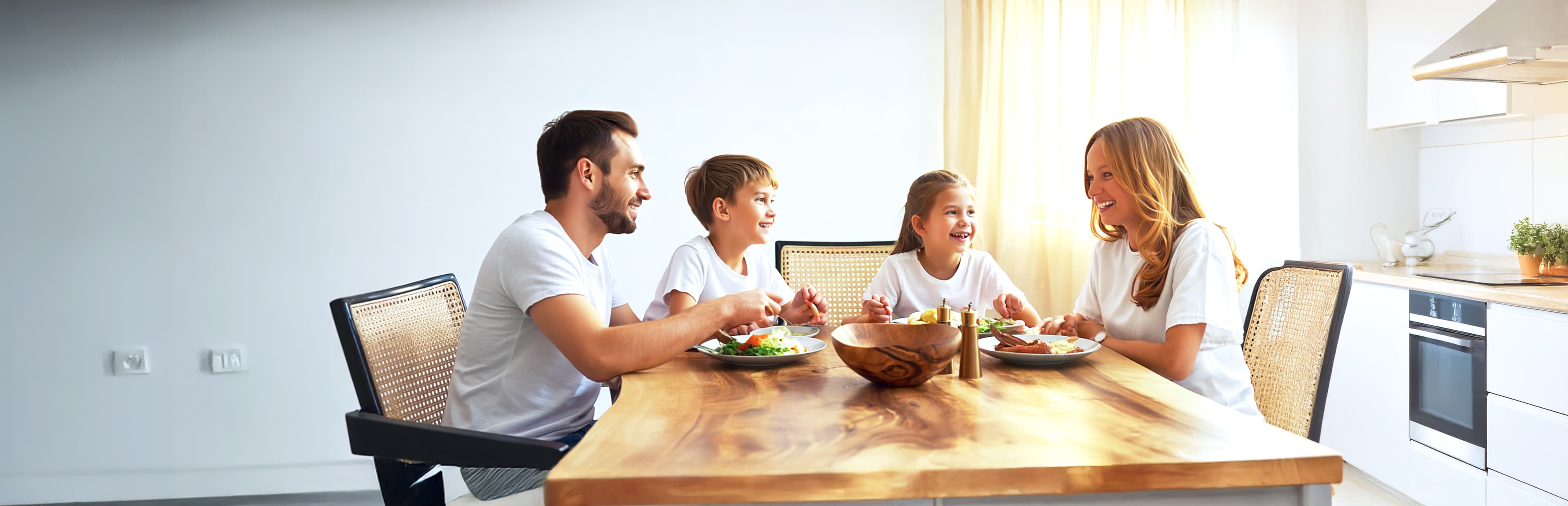 Eine vierköpfige Familie, zwei Erwachsene und zwei Kinder, sitzt lächelnd um einen hölzernen Esstisch und isst gemeinsam in einer hellen, modernen Küche eine Mahlzeit.