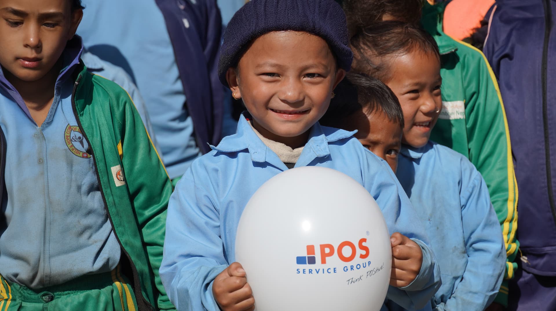 A smiling young child in a blue uniform and knit hat holds a white balloon with the POS Service Group logo. Other children in colorful uniforms stand beside him, all outdoors in sunlight.