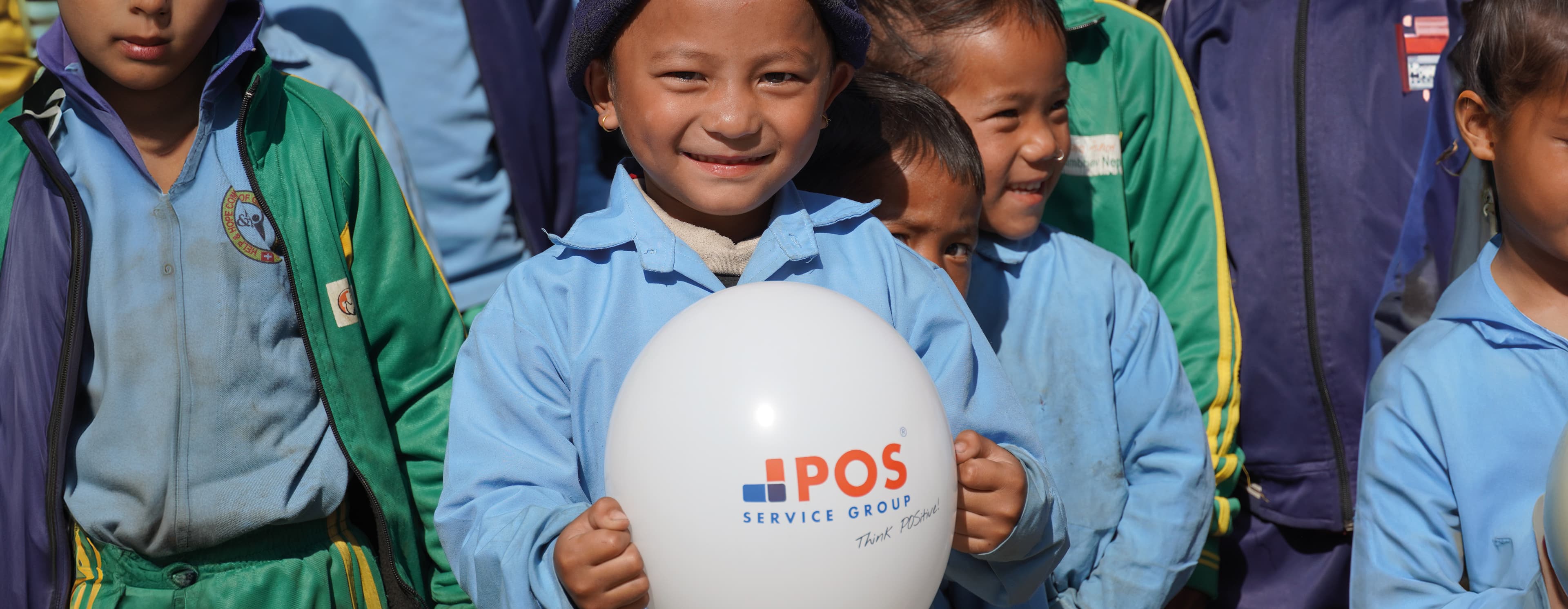 Een groep jonge kinderen in blauwe en groene schooluniformen staat buiten bij elkaar. Eén kind vooraan lacht terwijl het een witte ballon met het logo van POS Service Group vasthoudt.