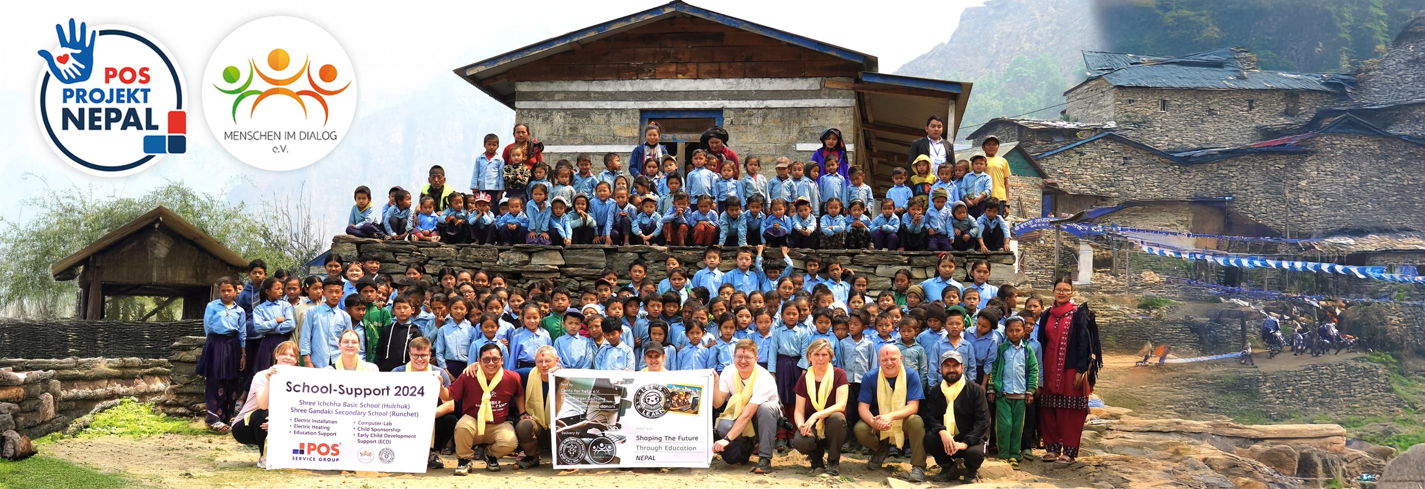 Een grote groep schoolkinderen en volwassenen poseert buiten voor stenen gebouwen en bergen, met spandoeken met logo's voor POS Projekt Nepal en MENSCH IM DIALOG e.V. en borden voor schoolondersteuning 2024.