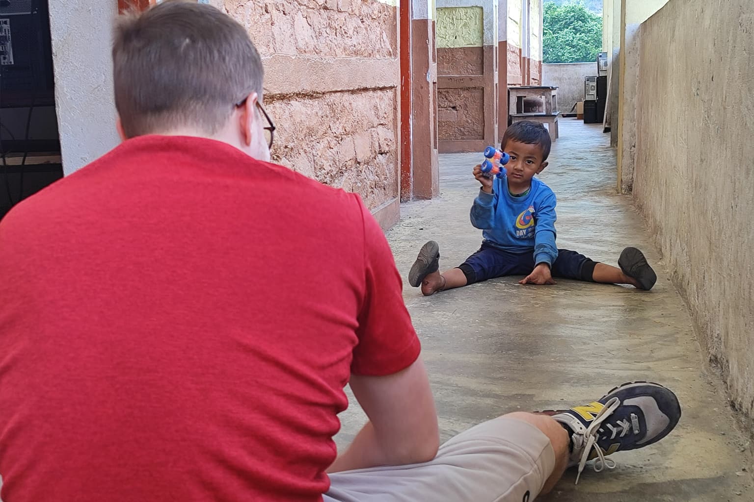 Een jongetje zit met gespreide benen op de grond, houdt een blauw speeltje omhoog en lacht naar een man in een rood shirt die tegenover hem zit in een gang met stenen muren.
