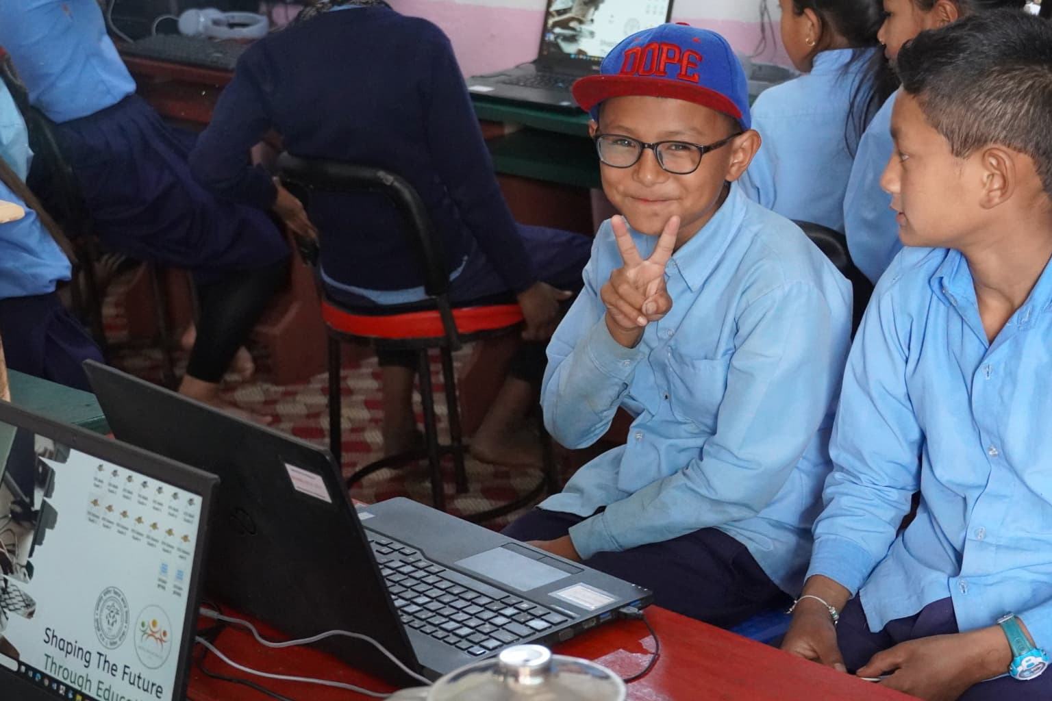 Een jongen met een blauw shirt en een rode pet zit naast een andere leerling, glimlacht en maakt een vredesteken voor een laptop in een klaslokaal, met andere leerlingen op de achtergrond.