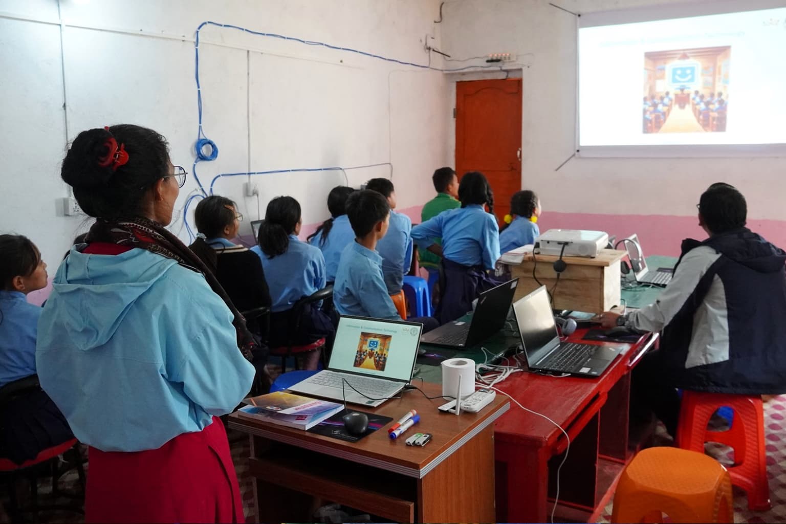 Een klaslokaal met leerlingen in blauwe uniformen, zittend aan bureaus, kijkend naar een presentatie op een scherm. Twee volwassenen houden toezicht terwijl laptops en lesmateriaal op de tafels staan. Het lokaal heeft witte muren en zichtbare bedrading.