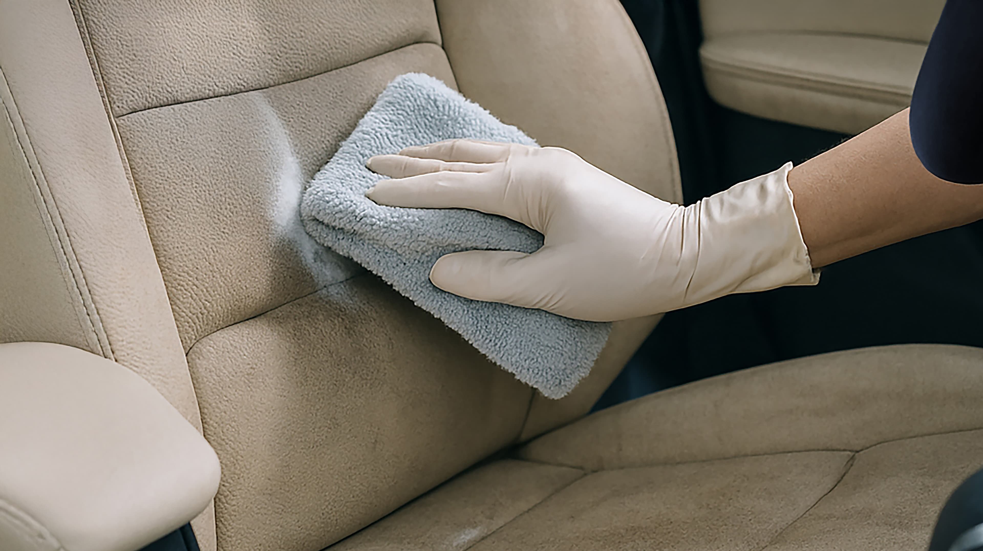 Une personne portant un gant en latex nettoie un siège de voiture beige avec un chiffon en microfibre bleu clair, en éliminant la saleté et les taches.