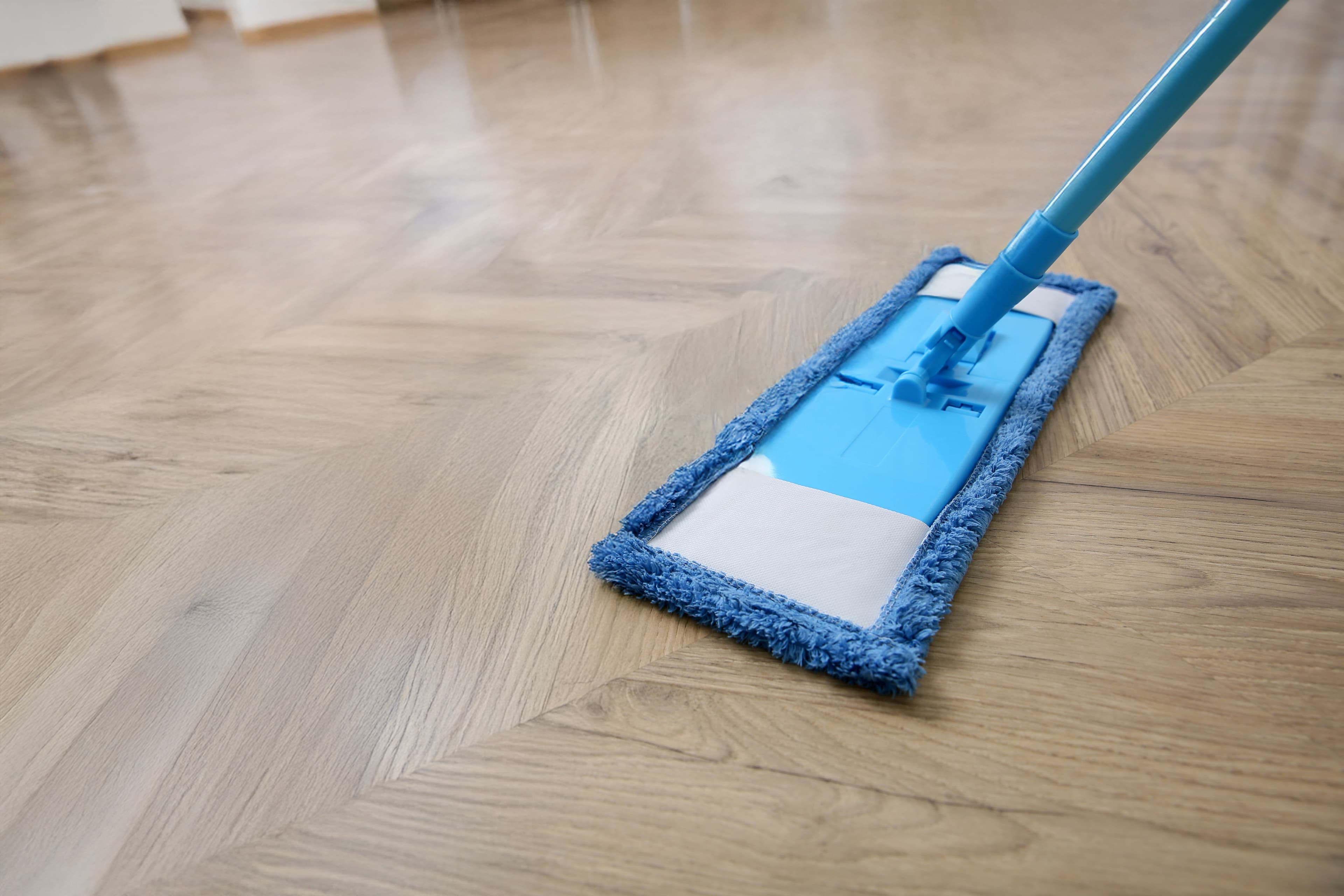 Nahaufnahme eines blauen Flachwischers, der einen hellen Holzboden mit Fischgrätenmuster wischt. Der Boden erscheint sauber und glänzend, und das natürliche Licht wird von der Oberfläche reflektiert.