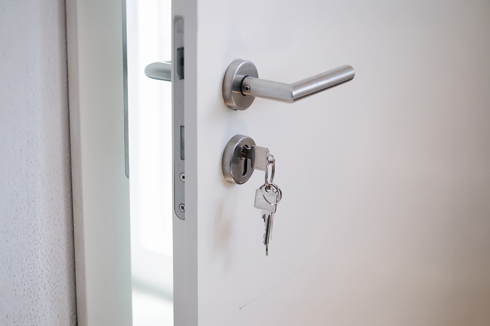 A white door is slightly open with a silver handle and a set of keys inserted in the lock. The keys have a keychain and are hanging from the door lock. Light shines through an opening in the background.