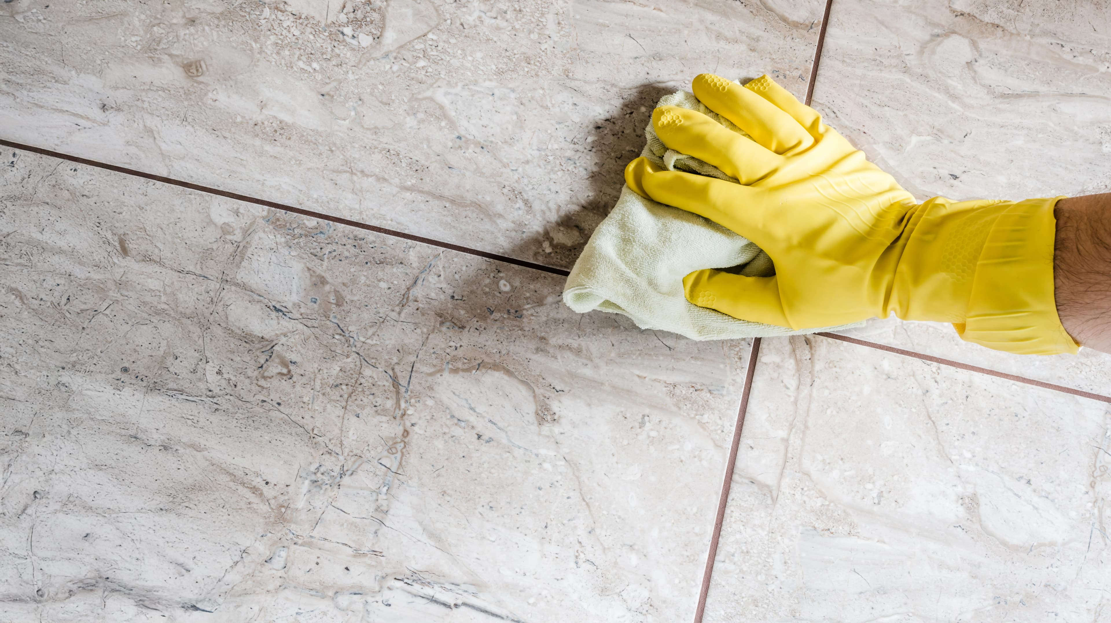 A person wearing a yellow rubber glove wipes a beige tiled floor with a white cloth, focusing on cleaning the grout lines between the tiles.