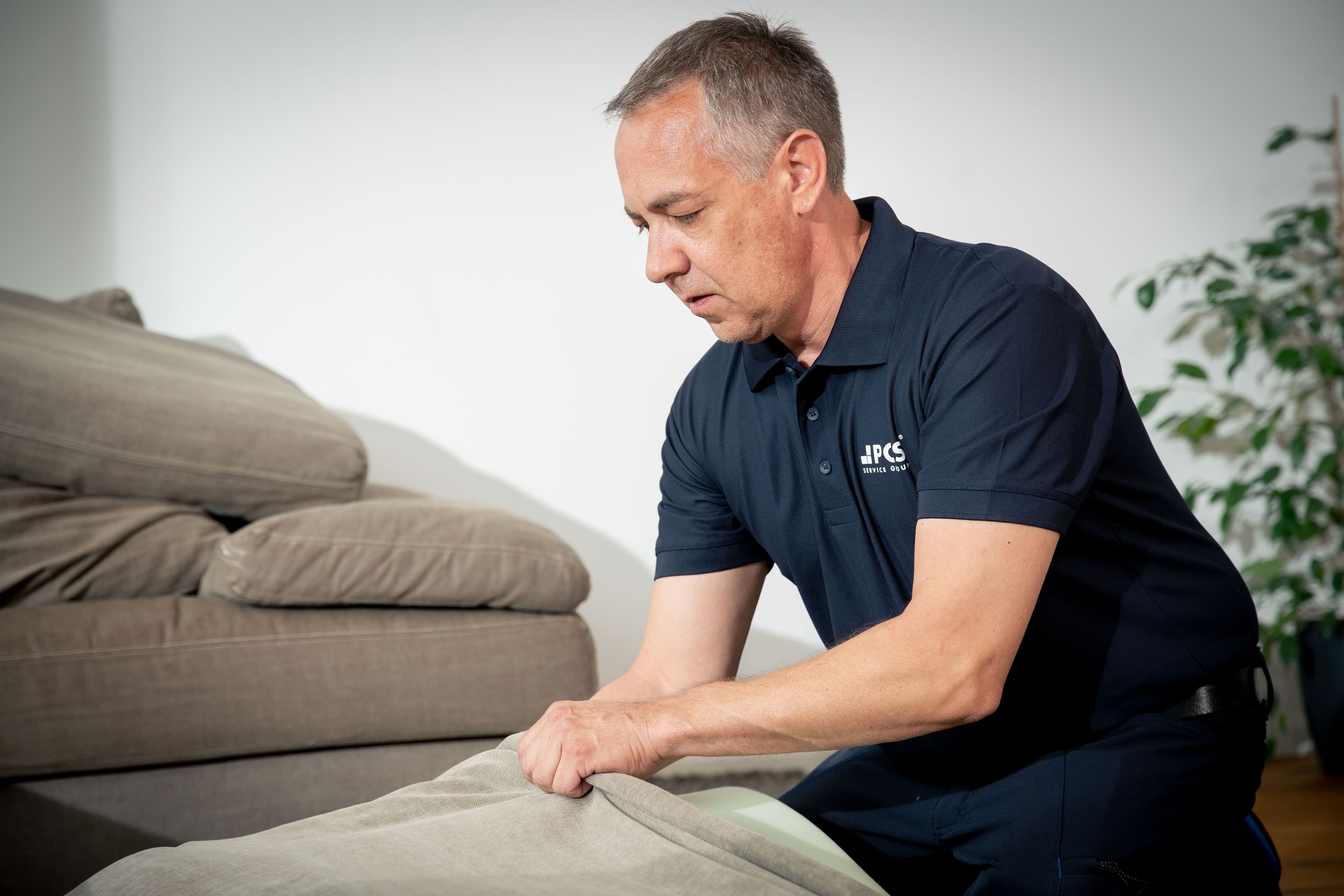 A man in a navy polo shirt removes a beige cushion cover from a sofa, with another couch and a plant visible in the background.