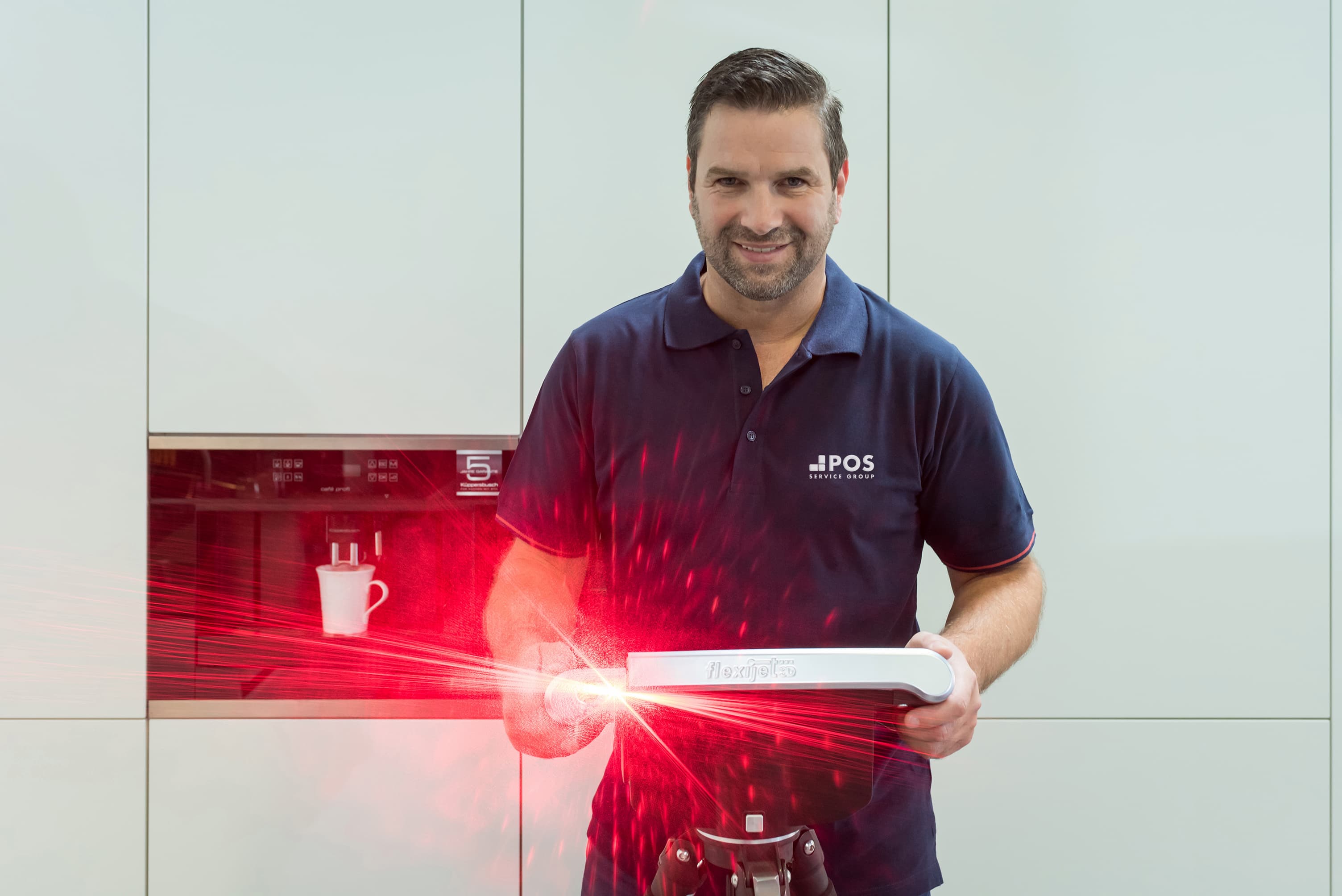 A man in a navy blue POS polo shirt smiles while holding a device emitting a bright red laser light in a modern kitchen, with a coffee cup and machine in the background.