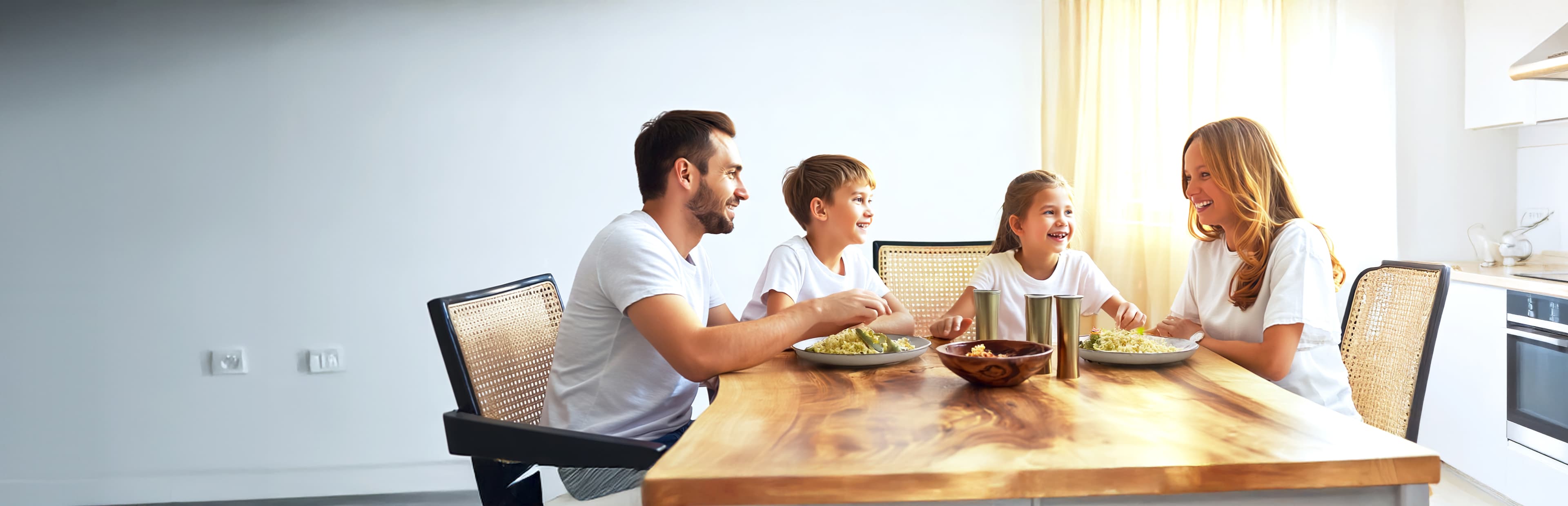 A family of four, two adults and two children, sit at a wooden dining table in a bright kitchen, smiling and enjoying a meal together.