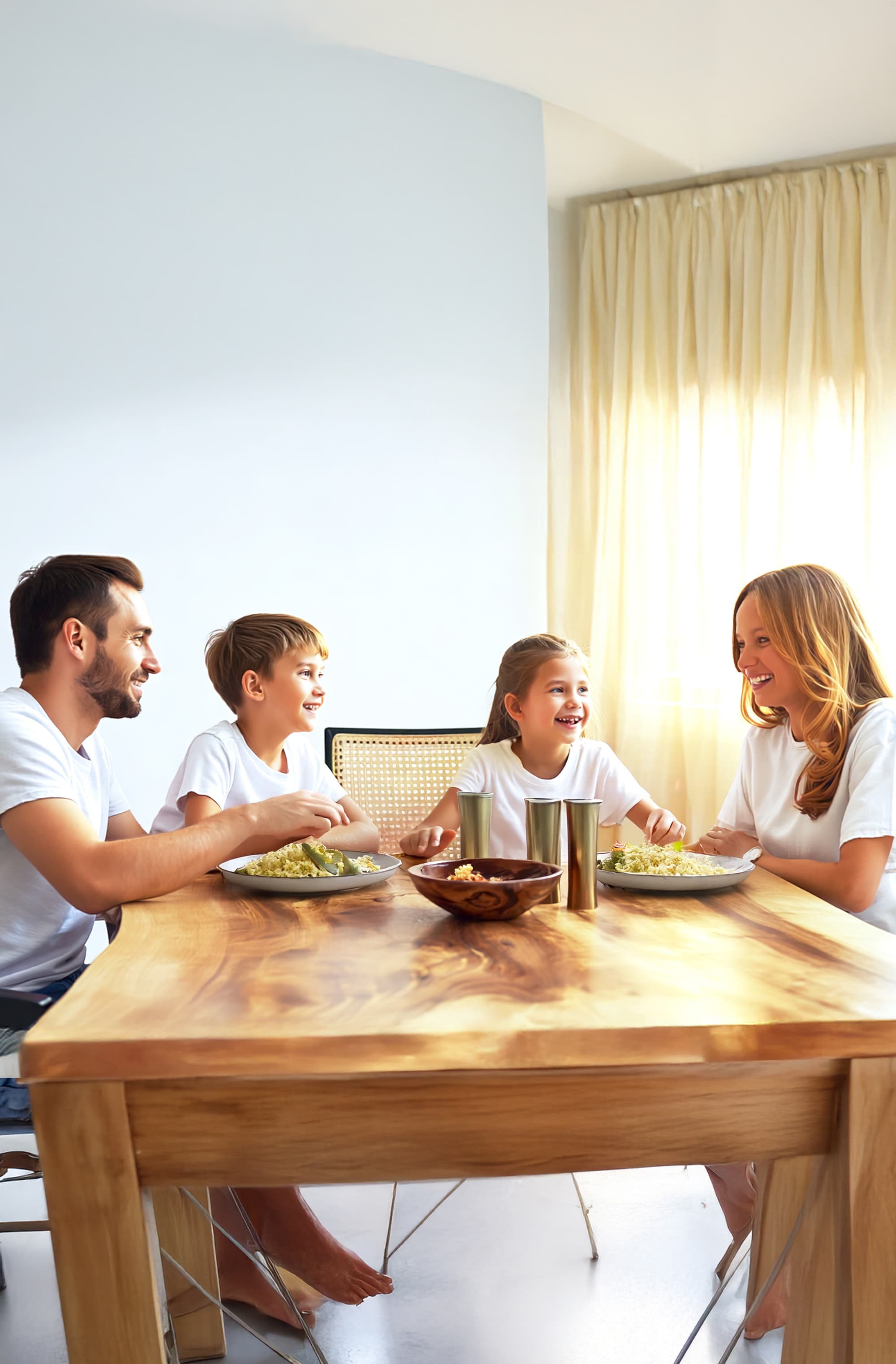 A family of four, dressed in white shirts, sits around a wooden dining table enjoying a meal together, smiling and talking in a bright room with sunlight streaming through curtains.